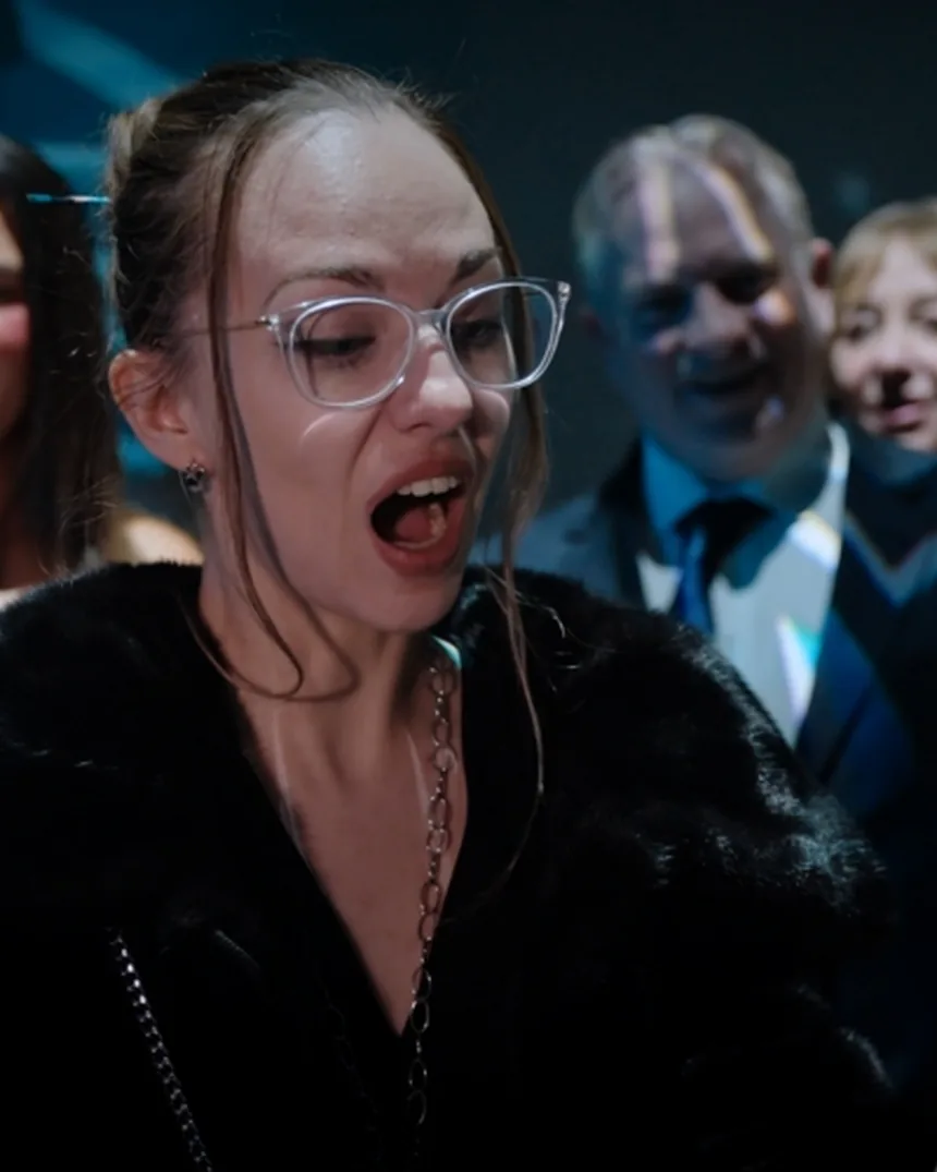 Guest in clear glasses and a black fur coat gasping at a magic trick while colleagues in suits watch behind her
