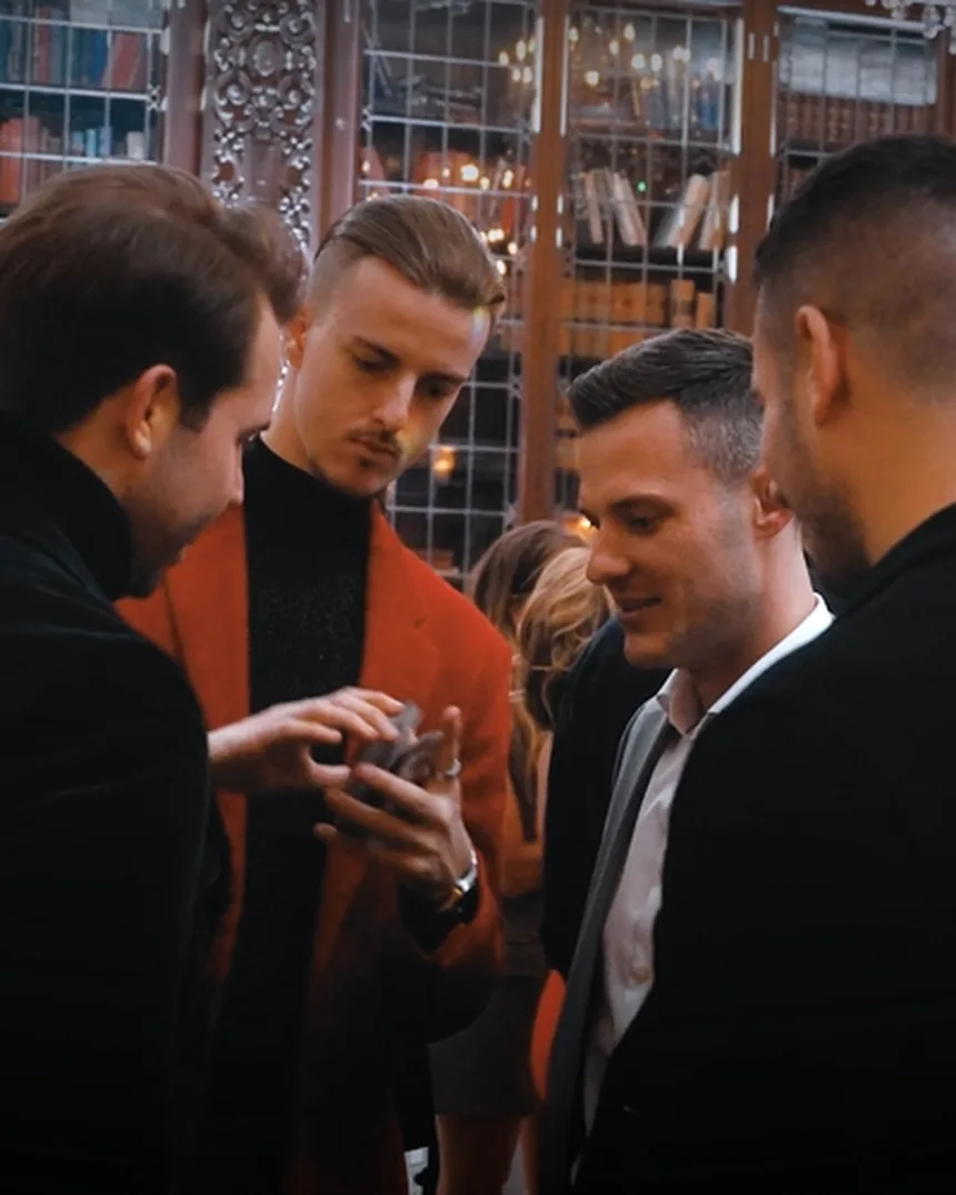 Roman Royss in a red jacket performing a card trick for a small group of guests in a library-style room