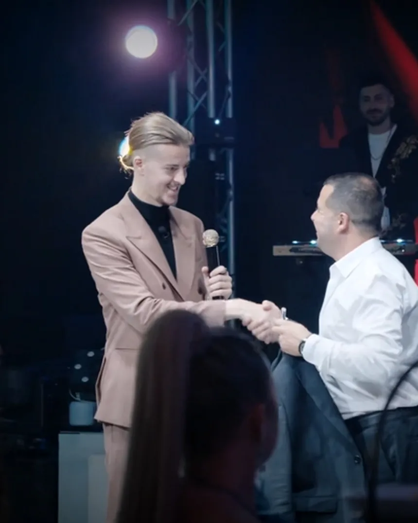 Roman Royss in a beige suit holding a microphone and shaking hands with a seated guest on stage