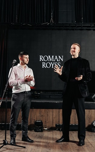 Roman on stage with a guest volunteer in front of his name on the screen