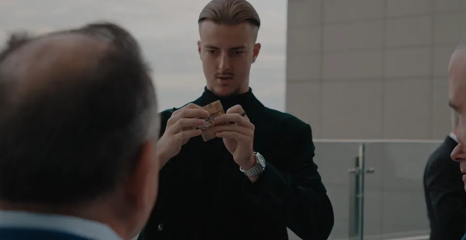 Roman performing a close-up trick with a small folded item for guests on an outdoor terrace