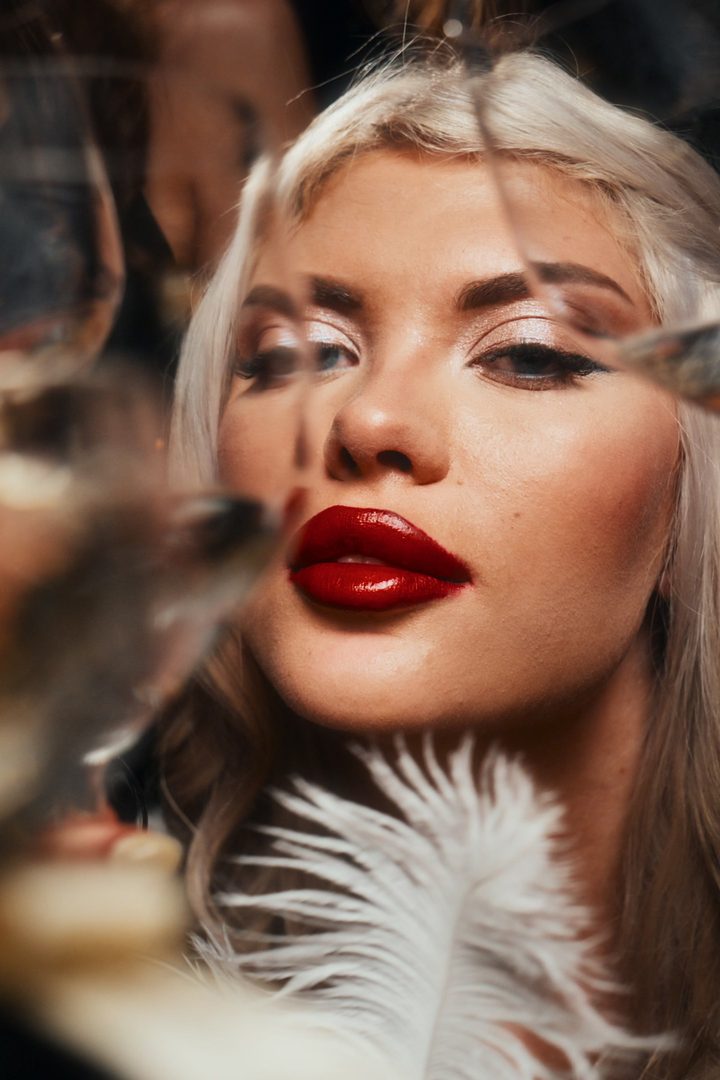 Close-up portrait of a woman with platinum hair and red lipstick seen through wine glasses, a white feather at her chin
