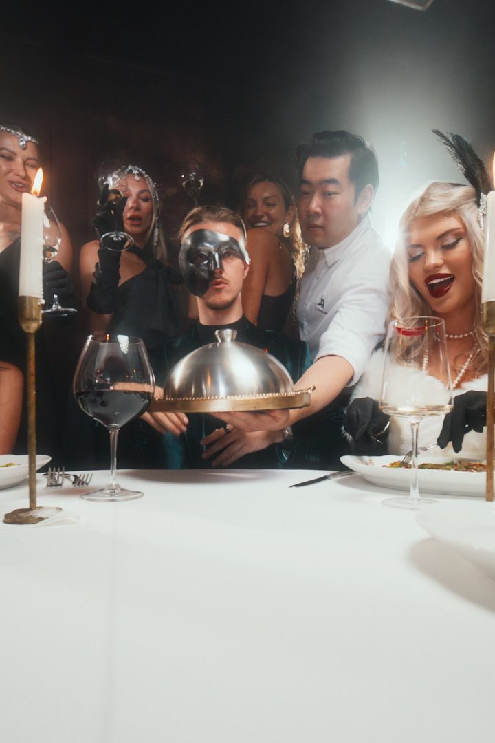 Man in a black half mask at a candlelit table with a silver cloche, guests in 1920s dress behind him