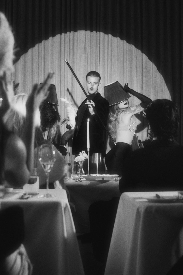 Black and white scene of a man holding a cane before a draped curtain, a woman in a lampshade hat beside him