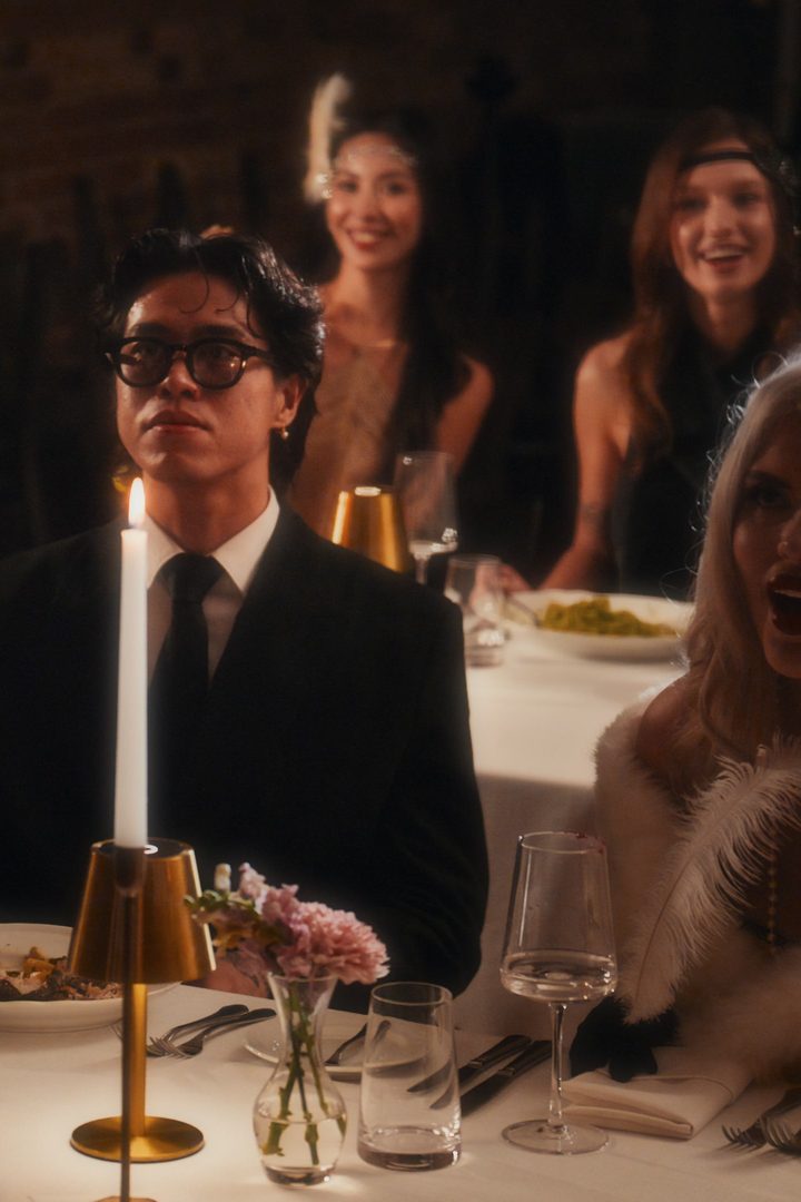 Man in glasses and a black suit beside a lit candle at a dinner table, smiling guests in 1920s style behind him