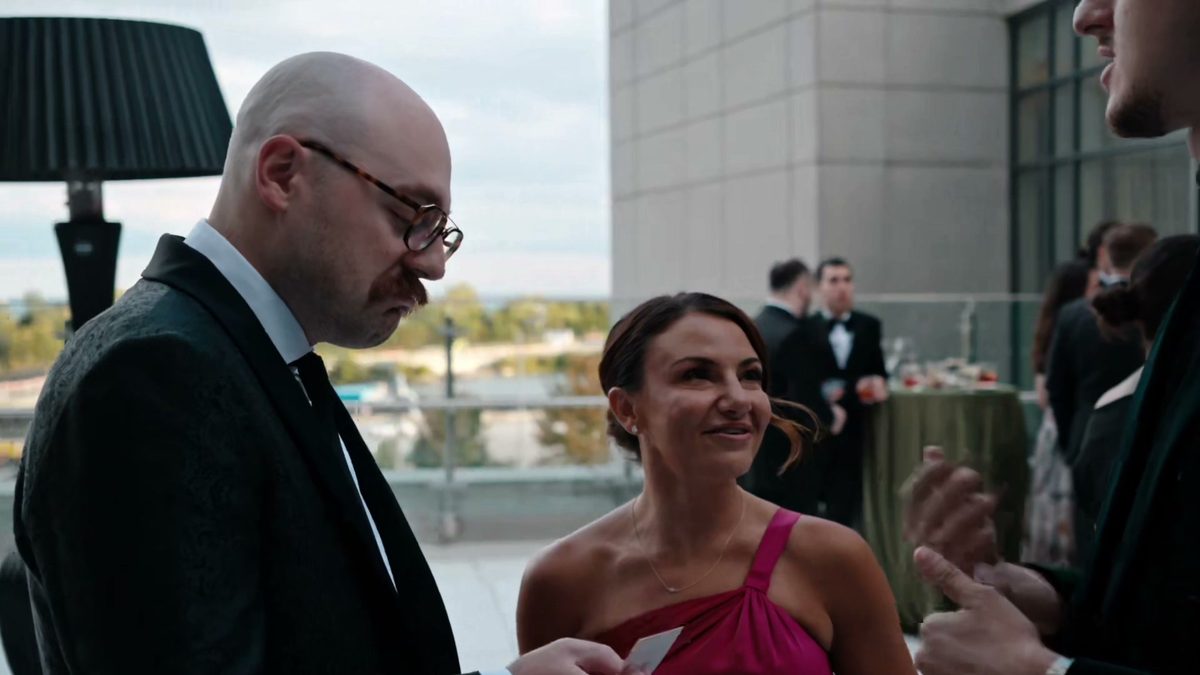 A guest in a pink dress smiling at a playing card beside a man in glasses on a terrace