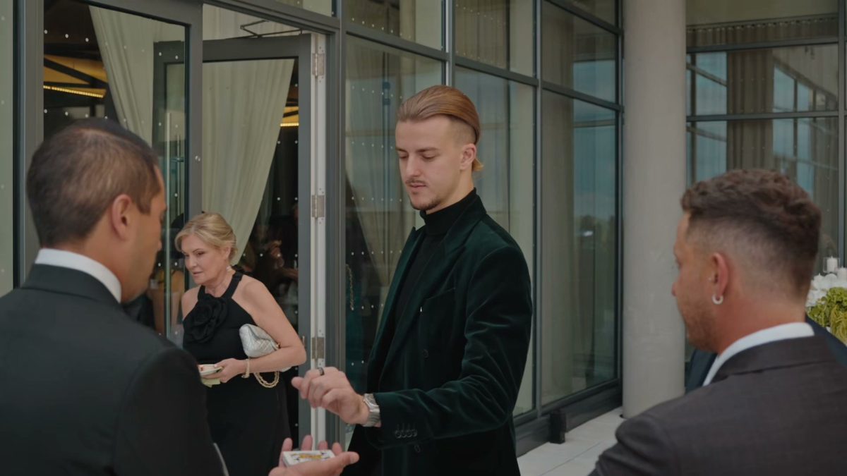 Roman Royss in a green velvet jacket performing close-up magic for guests on a terrace by glass doors