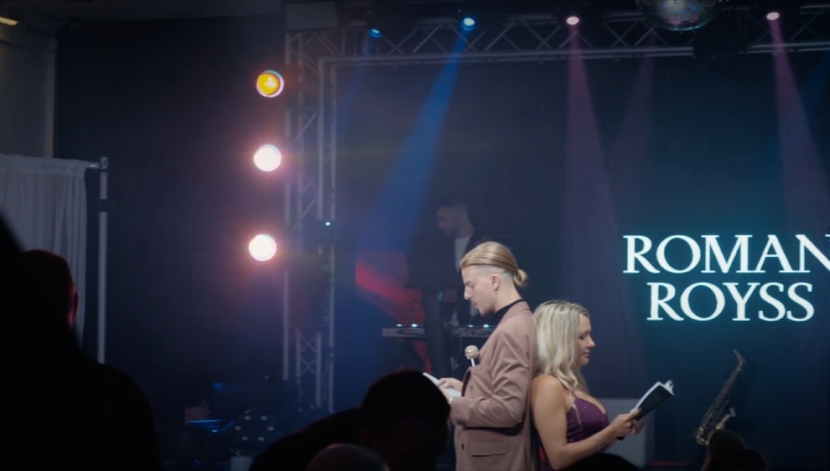 Roman Royss on a lit stage beside a guest reading a card, his name in lights behind them
