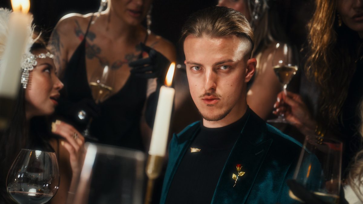 Roman Royss in a teal velvet jacket at a candlelit dinner table, guests behind him