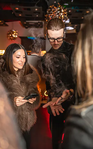 Roman Royss in glasses lets a wisp of smoke rise from his hands while a woman in a fur coat watches at a brand launch