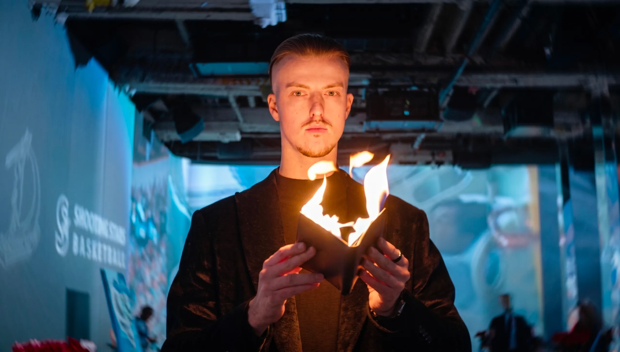 Roman Royss in a black velvet blazer holds a small dark object on fire in his hand, under a blue-lit industrial ceiling