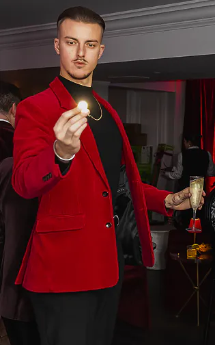 Roman Royss in a red velvet blazer holds up a small glowing object between his fingertips at a holiday event
